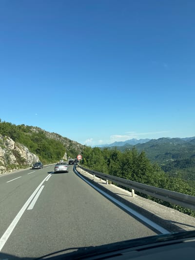 The image depicts a road in Dobrsko selo, Montenegro. The road is asphalt and has two white lines marking the lanes. There are several cars on the road, including a black car in the foreground and other vehicles further ahead. A guardrail runs along the side of the road, separating it from a steep, rocky slope covered in green vegetation. In the background, there are mountains covered in trees. The sky is clear and blue. There is a no-entry sign visible further along the road. The scene appears to be taken from inside a vehicle.