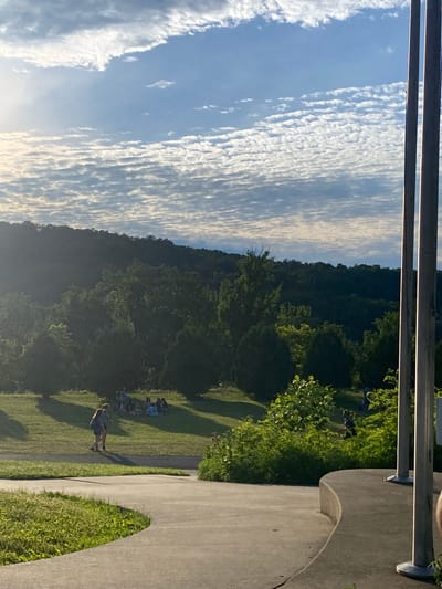 The image depicts an outdoor scene with a gentle slope in the foreground leading to a grassy area. Two individuals are walking on a paved path in the midground, casting long shadows. In the background, a group of people are gathered on the grass, some sitting and others standing. Beyond them, a dense forest covers a rolling hill, with a bright sky filled with scattered clouds above. The overall atmosphere is serene and appears to be a park or recreational area. The location is identified as Washington Township, United States of America.