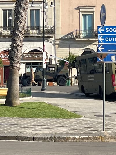The image depicts a street scene in Galatina, Italy. Three military vehicles are parked along the street, in front of a building housing the "Caffe Alighieri" and "Gelateria Artigianale." A palm tree is visible to the left. A street sign points towards "Sogliano" and "Cutrofiano." A narrow strip of green grass separates the sidewalk from the road. The buildings have windows with white trim.