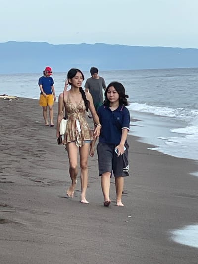 The image depicts a beach scene in Dauin, Philippines. In the foreground, two young females are walking along the dark sand, with the ocean and waves on their right. The female on the left wears a patterned, sleeveless dress and is carrying a white bag and a phone. The female on the right is wearing a dark blue polo shirt with white trim and dark shorts, and she is also holding a phone. In the middle ground, a male wearing a gray t-shirt and a person in a blue t-shirt and yellow shorts are walking further down the beach. In the background, a hazy mountain range is visible against a light sky.