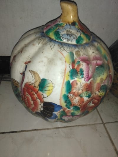The image displays a single, lidded ceramic vessel, bulbous and fluted in shape, resembling a pumpkin or gourd. The object features a predominantly off-white base with extensive polychrome enamel decoration, including vibrant floral motifs in red, orange, pink, yellow, blue, and green. A stylized bird, appearing to be a peacock, with red plumage, a brown body, and blue and black tail feathers, is depicted on one side. The lid is adorned with blue floral patterns and has a short, light brown finial. The vessel's surface exhibits dust and grime. It rests on a light-colored tiled floor with dark grout lines. The background consists of a plain, light-colored wall, also appearing somewhat textured or dirty. No human figures or activities are present. The environmental setting is an interior space. The precise location within Ciudad de San José, Costa Rica, is not discernible from the visual evidence presented in the image.