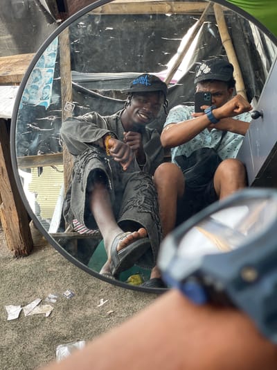 Two young Black men are captured in a mirror selfie. The man on the left, wearing a dark grey distressed jacket and pants, is smiling at the camera. He has braided hair and is wearing a black baseball cap with a blue emblem and a gold beaded bracelet on his left wrist. The man on the right, wearing a light blue patterned shirt and black shorts, is holding a smartphone to take the picture and is looking intently at the screen. He is wearing a black baseball cap with white floral embroidery and a dark blue wristwatch. They are seated closely together, appearing to be outdoors in a rustic environment. The background visible in the mirror shows rough wooden structures and what appears to be a tarp or sheeting. The ground beneath them is covered in sand and debris, with some scattered papers. This scene is observed in the city of Ojomu, Nigeria.