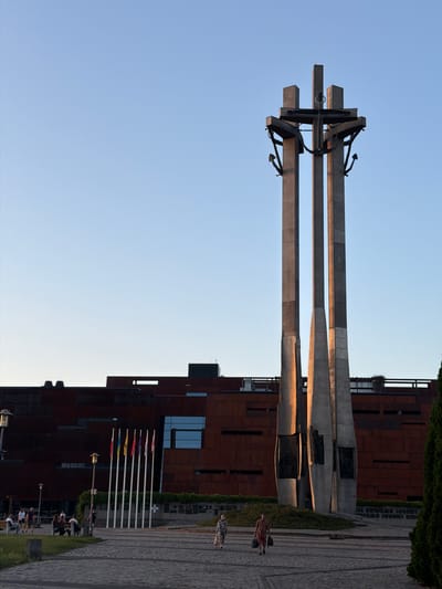 This image captures an outdoor scene in Gdańsk, Poland, featuring a monument with three large, horizontal concrete beams. The monument is adorned with black anchor elements. To the right of the monument, a modern building with a rusted metal facade stands. In front of the building, a paved walkway is lined with flags on poles and green bushes. Several people are visible, some walking along the paved area, while others are seated on benches. One person in the foreground is wearing a checkered shirt and shorts, and another in the background is wearing a plaid shirt. The lighting suggests it is either early morning or late afternoon, with the sun casting warm light on the monument's beams. The sky is clear and blue, indicating fair weather.
