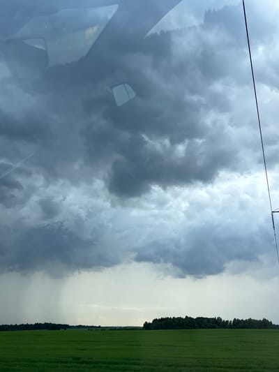 A wide shot depicts a stormy sky over a rural landscape. In the foreground, a vibrant green field stretches horizontally. A dense, dark green tree line forms a distant border separating the field from the sky. The sky is filled with dramatic, dark gray storm clouds. On the left side of the frame, a visible rain shower is falling, creating a lighter gray vertical shaft. The right side of the frame shows a thin black wire extending vertically from top to bottom. Reflections of what appear to be vehicle interior elements, possibly the windshield and dashboard, are visible in the upper portion of the image, suggesting the photo was taken from inside a vehicle. The scene is located in or near Поварёнки, Russia.