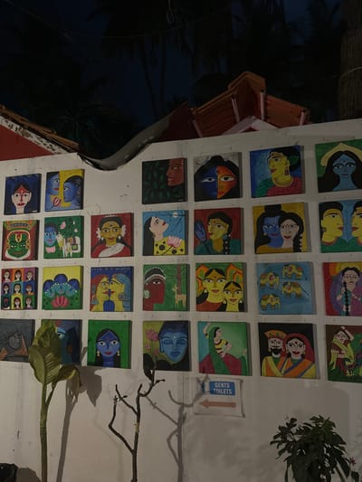 The image depicts an illuminated outdoor wall adorned with approximately 36 square, colorful folk-art paintings, likely in the Madhubani style, located in Kolamb, India. The artwork, arranged in roughly six rows and six columns, primarily features stylized depictions of human faces, couples (including divine figures like Radha and Krishna), animals (elephants, deer), and natural elements such as flowers and leaves. The dominant color palette includes blues, reds, yellows, and greens.

Above the white wall, terracotta-tiled rooftops are visible, and dark silhouettes of palm trees are present against a dark, possibly night sky, indicating an evening or nighttime setting with artificial light source illuminating the wall. In the lower right section of the wall, a white rectangular sign with a blue border reads "GENTS TOILETS" and includes a male pictogram, with an arrow pointing to the right. To the left of this sign, a tall plant with large green leaves stands, casting a shadow on the wall, and a smaller, bare branch-like plant is also present. Another leafy plant is visible in the bottom right corner. No people are visible engaging directly with the artwork or in the immediate foreground, and no direct actions or interactions are depicted outside of the artwork itself.