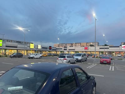 The image shows a parking lot in Giurgiu, Romania, in front of a shopping complex with various storefronts. The sky is a mix of blue and gray, suggesting either dusk or dawn. The parking lot is filled with several cars of various makes and models. In the foreground, a blue car is partially visible, obscuring some of the view. There are streetlights illuminated throughout the parking lot. The storefronts in the background have signs for shops like TAKKO Fashion, kik, and New Yorker.