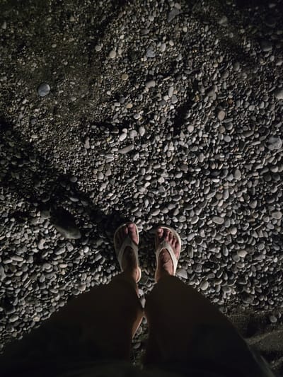 The image shows a top-down view of a person's feet wearing white flip-flops, standing on a surface covered in small, smooth, gray and brown pebbles. The lighting suggests it is nighttime, with strong shadows cast by the person's legs and feet onto the pebble ground. The scene appears to be an outdoor area, possibly a beach or a path, given the texture of the ground. No specific landmarks or indicators of Salerno, Italy, are visible in the image itself.