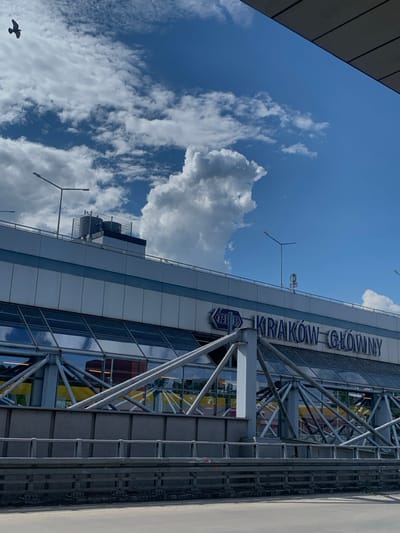 The image shows a view of the Kraków Główny train station in Kraków, Poland, at 50.068 latitude, 19.949 longitude. The image is taken from a distance and shows the station's exterior, with the "Kraków Główny" sign visible on the front of the building. The station is a large, modern building with a steel and glass facade. The top of the building is visible, and the sign is located on the upper level of the structure. The building is situated behind a tall, gray metal fence with a black railing on top. The image is taken on a clear day with a few clouds in the blue sky, and a single bird is flying in the upper left corner of the image. There are several streetlights on the roof of the building, including a tall, white streetlamp with a square base in the top right corner of the building. No people are visible.