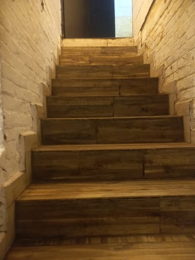 A dimly lit space features a distinctive wooden staircase or tiered structure. Vertical wooden planks line one side, potentially forming a wall or support system. The floor is made of lighter-colored, possibly stone or concrete blocks laid horizontally. A window or doorway is visible in the background, providing a glimpse of a lighter area. The composition emphasizes the architectural details and textural contrast between the vertical wooden elements and the horizontal flooring.