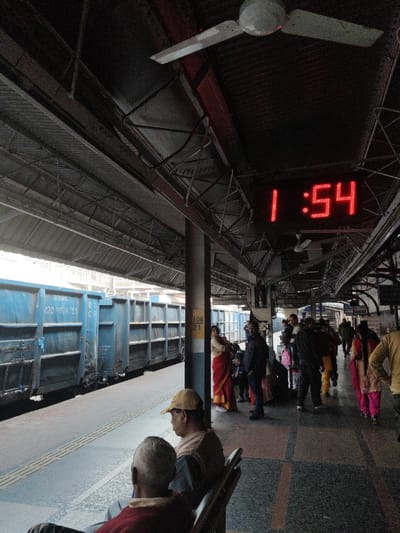 The image depicts a train station platform in Bardhaman, India. The scene is illuminated by daylight from the left, with the overhead platform roof casting shadows across the foreground and midground. A prominent red digital clock mounted overhead displays "1:54". Multiple white ceiling fans are suspended from the dark metal framework of the roof structure.
To the left, a blue freight train consisting of multiple boxcar-type wagons is stationary alongside the platform. Markings such as "BOXNHL" and "2201924755" are visible on the side of the nearest wagon. The platform surface is tiled in a pattern of alternating dark and lighter greyish-blue rectangular sections, with a raised tactile strip running along the edge adjacent to the tracks.
In the immediate foreground, two older men are seated on a bench with their backs to the viewer. The man closer to the left wears a yellow cap and a light-colored vest over a dark shirt. The man to his right wears a red top.
In the midground, a light grey support column with a yellow and white marker displaying "106 21" stands vertically. A woman in a red saree leans against this column, holding a bundled infant. To her right, a group of approximately eight to ten individuals are standing and walking on the platform. These individuals are dressed in varied attire, including dark jackets, sarees (one pink, one yellow visible), and casual clothing. Several children are interspersed within this group. People appear to be waiting or moving along the platform.