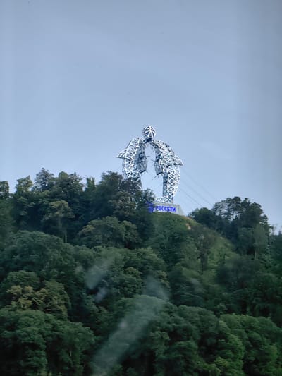 This media file depicts a large, illuminated sculpture atop a verdant, tree-covered hill. The sculpture, made of metal lattice, appears to be two abstract figures or shapes leaning towards each other, forming an arch. The top of the sculpture has a spherical element. The sculpture is lit with blue lights, and the word "РОССЕТИ" (ROSSETI) is visible in blue illuminated letters at the base of the sculpture.

The scene is set in a natural environment, with the hill densely covered in green trees. The sky above is a pale, overcast blue, suggesting either early morning, late afternoon, or a cloudy day. The overall impression is one of a public art installation or monument situated in a scenic, possibly mountainous or hilly, location. The presence of what appears to be power lines or cables extending from the sculpture further suggests a connection to infrastructure or a viewing point.

The time of day is likely either dawn or dusk due to the lighting on the sculpture and the overcast sky. The weather appears to be mild, with no strong indications of rain or extreme conditions. The location is identified as Vysokoye, Russia, which provides geographical context for this monument. The media file captures a static view of this impressive structure against a natural backdrop.