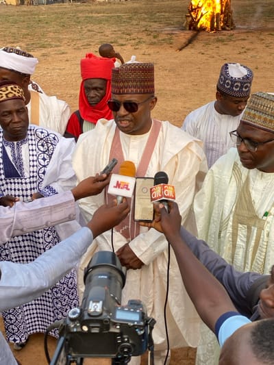 The image captures an outdoor press engagement or public address involving a central figure surrounded by media and other individuals. A man, dressed in a light-colored traditional robe, a patterned cap, and sunglasses, is the primary subject, appearing to speak to reporters with a slight smile. He is holding a smartphone, possibly for recording, and several microphones are extended towards him. Two microphones prominently feature the "ait" logo, while another bears the "NTA" logo.
To the right, a person operates a professional camera, identified as a Canon, aimed at the interviewee. Other men in various traditional attire, including patterned caps and robes, are visible around the central figure, observing or participating. One individual with a red head covering is partially seen behind the main speaker.
The setting is an open, unpaved area, likely outdoors during the daytime, given the natural light and the presence of sunglasses. In the background, a large bonfire is actively burning, adding a dramatic element to the scene. The attire and the media presence suggest a public event or announcement taking place, consistent with a location like Agunu, Nigeria. The overall atmosphere appears engaged and active, indicative of a newsworthy occasion.