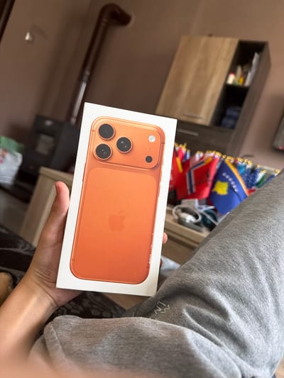 A person is holding an iPhone 12 box, which displays a picture of an orange iPhone 12. The background is blurry, but it appears to be an indoor setting, possibly a living room or bedroom, with a cabinet and a piece of furniture visible. The person is wearing grey sweatpants. The image was taken in Peshtera, Bulgaria.