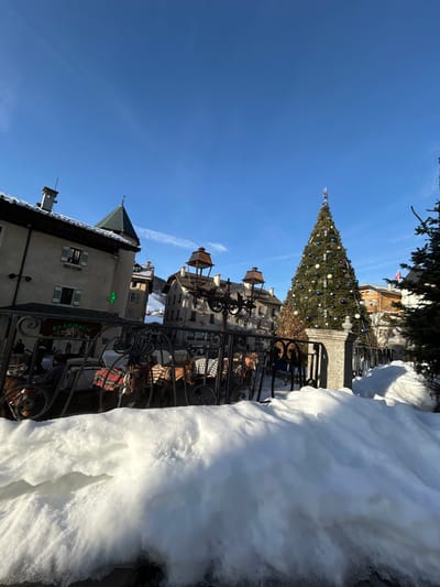 A winter scene captures a European town square adorned with a tall Christmas tree. Buildings with distinct architectural styles, including one marked "Pharmacie," are visible. A wrought-iron fence stands between the viewer and the town. Large snowdrifts line the edge of the scene, adding to the wintry atmosphere. The sky is a clear blue. Lanterns are hanging over the fence and buildings.