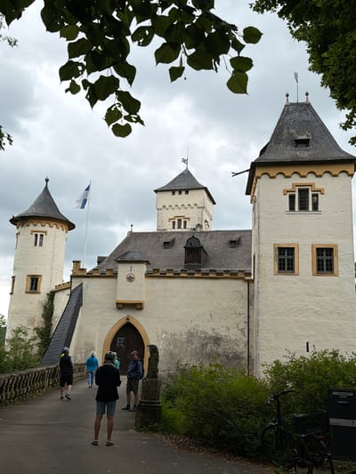 The image shows a cloudy day at the Heiligenstein Castle in Heiligenstadt in. OFr., Germany. A group of people, some wearing helmets, are walking on a path leading to the castle's entrance. The castle itself is a cream-colored building with several towers, a prominent flag flying from a flagpole, and dark-tiled roofs. The foreground includes green foliage and a parked bicycle.