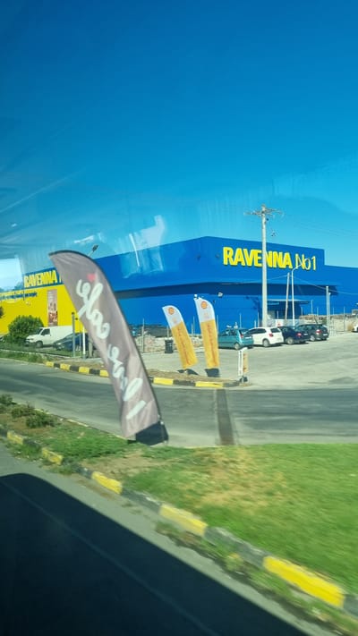 The image depicts the exterior of a "RAVENNA No1" building, a large blue structure with yellow lettering, located in Larissa, Greece. In front of the building, there is a parking lot with several cars. Two yellow flag banners, possibly advertising Shell, stand near the entrance. A gray flag banner with white writing is prominently displayed in the foreground, partially obscuring the view of the building. The area around the building features green grassy areas and a paved road with yellow and black striped curbs. The sky above is a clear, bright blue. The scene appears to be captured from a moving vehicle, as indicated by the slight blur and reflection on the window.