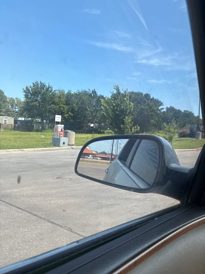 The image shows a view from inside a vehicle, looking out the side window and at the side mirror. The side mirror reflects a portion of the car's interior, a portion of the road, and some buildings in the distance. Outside the car, there is a gas station with a "7-Eleven Air" sign. Trees and grass are visible in the background, along with some residential structures. The sky is blue with some wispy clouds. The location is Balch Springs, United States of America.