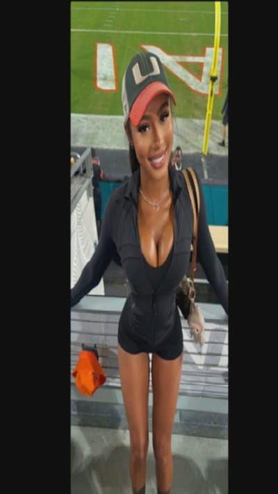 A woman wearing a black athletic outfit and a "U" logo baseball cap stands in stadium bleachers. The cap has a green bill and a mesh back. She is fair-skinned with dark hair. Her necklace is visible. Behind her, a football field with yard lines and a "U" emblem is seen. Other people and stadium structures are also visible in the background. An orange cap sits on the bleachers to her left.