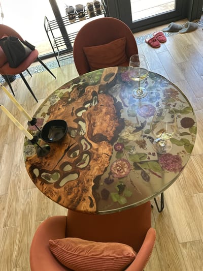 A high-angle shot shows a round table with a distinctive design, surrounded by chairs, likely indoors in Малеевка, Russia. The tabletop features a combination of wood and floral patterns, with a clear resin-like material encasing dried flowers and leaves around a central section of figured wood with irregular light-colored inlays. Several wine glasses are placed on the table's surface. A small, square-shaped black bowl sits at the table's edge, along with two tall, thin, light-colored candleholders topped with black bases.
Two upholstered armchairs are visible, one on either side of the table, in a muted orange-brown hue. One chair has a matching pillow. In the background, to the left, is a shoe rack holding several pairs of shoes. The floor appears to be tiled with light-colored wood-patterned tiles, except for a small area near the door showing patterned tilework. Slippers hang on the wall next to the doorframe. The lighting seems to be natural, casting soft shadows across the scene.