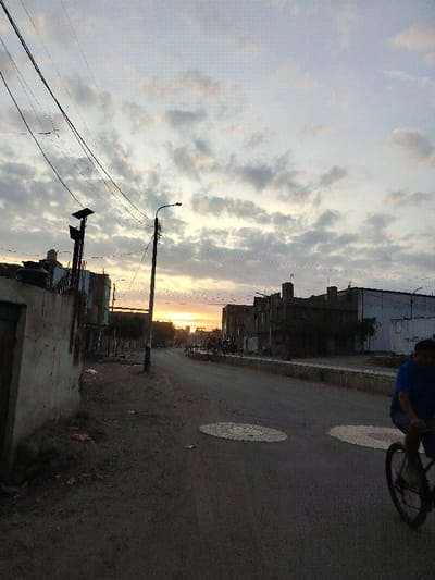 The image displays an urban street scene in Florencia de Mora, Peru, captured during either sunrise or sunset, indicated by the low sun casting an orange glow on the horizon and the generally dim lighting. The sky features scattered clouds.
In the left foreground, a weathered concrete wall borders an unpaved area strewn with debris. Behind the wall, utility poles rise, supporting multiple overhead power or communication lines that extend across the frame. One pole is equipped with what appears to be a solar panel. Rooftops and portions of multi-story buildings, including a black water tank, are visible behind the wall.
The central and right portions of the image are dominated by an asphalt road, partially shadowed. Two circular, light-colored patches are distinctly visible on the road surface in the midground. A low concrete barrier runs along the right side of the street, separating it from a series of multi-story buildings, which exhibit various architectural styles and states of repair.
In the lower right foreground, a lone individual wearing a blue shirt is visible, riding a bicycle away from the viewer. Only the torso, right arm, and the bicycle's front wheel and handlebars are clearly discernible. Distant buildings are silhouetted against the bright horizon.
