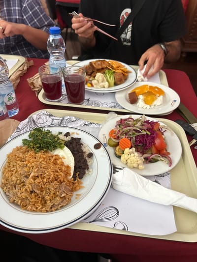 This image captures a close-up view of a meal being served on a tray. Several plates of food are visible, along with glasses of red juice and a bottle of water. One plate contains a salad with tomatoes, red cabbage, onions, cauliflower, and pickles. Another plate features rice, black beans, a fried egg, green vegetables, and shredded meat. A smaller plate holds what appears to be dessert with fruit and cheese. A person's hands are seen reaching for the food, suggesting the meal is in progress. The setting appears to be an indoor dining area, possibly a restaurant, with a red tablecloth. The time of day is not explicitly indicated, but the lighting suggests daytime. The overall impression is one of a casual meal. Visible text includes "DUNAS" on some of the plates, and "UP" and a barcode on the water bottle.