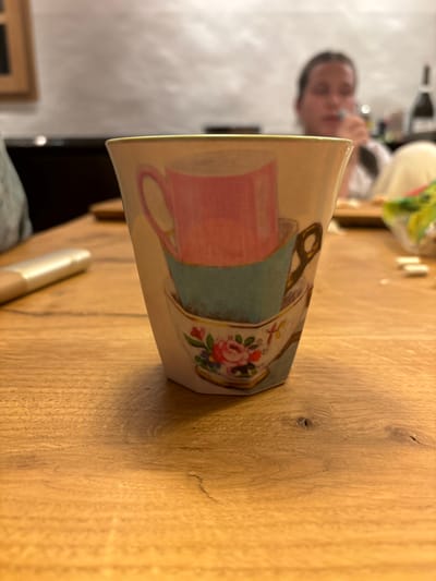 A white ceramic cup, decorated with three stacked teacups, sits on a wooden table in Celerina/Schlarigna, Switzerland. The teacups are pink, blue and white with a floral pattern. The top teacup is pink and the bottom teacup has a pink rose on it. The handle of the top teacup is visible. The cup is the focus of the photo. A blurred figure is seated in the background, seemingly looking at the cup. The table is light brown wood with visible grain and a natural texture. The background is blurry. The room is dimly lit. There is a sliver of a white object behind the person in the background. The photo is taken from a slightly above view of the cup.  The coordinates (latitude: 46.514, longitude: 9.859) correspond to the village of Celerina/Schlarigna, Switzerland.  The cup is in the center of the image, and the wooden table is the foreground.