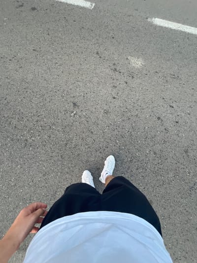 The image shows a first-person perspective of someone walking on asphalt pavement. The person is wearing black shorts, a white t-shirt, and white sneakers. A hand is visible on the left side of the frame. There are two white painted lines on the asphalt, indicating a road marking. The scene appears to be outdoors, possibly in a street or pathway. The location is identified as Vladimirovca, Moldova.