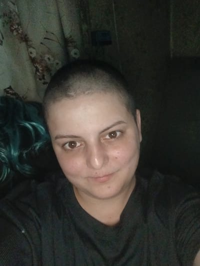 The image is a close-up, rotated portrait of a person with a shaved head and teal-colored hair peeking from the top of the frame. The subject is looking directly at the camera with a neutral expression. Their skin is fair, and they have brown eyes. Freckles are visible on their cheeks and nose. They are wearing a dark-colored, possibly black, crew-neck shirt.
The background appears to be a textured surface with a floral pattern in muted green and brown tones, possibly a sofa or a blanket. The lighting is dim, suggesting the photo was taken indoors in low light conditions. The overall mood is intimate and personal, as it seems to be a selfie. No text is visible in the image. The given location context is Chutove, Ukraine.