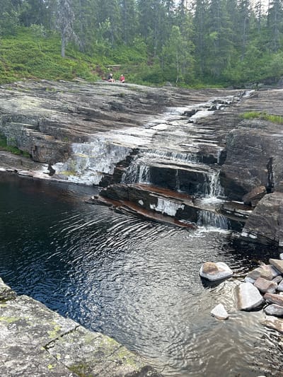 The image shows a natural scene with a rocky cliff face and a body of water. The cliff is layered and appears wet in places, with streaks of white water cascading down. The water is dark and rippling, with some rocks visible on the surface near the edge of the cliff. In the background, a dense forest of green trees covers a slope.
There are three people visible on the side of the cliff, near the treeline. They appear to be engaged in some activity on the rocky slope. One person is wearing a red top, another a dark top, and a third is partially obscured. They are positioned on a slightly less steep section of the cliff, with vegetation growing around them.
The lighting suggests it is daytime, possibly late afternoon given the shadows and the illumination on the water. The weather appears clear. The overall impression is one of a serene natural landscape, possibly a hiking or outdoor recreational area in Norway. There is no visible text in the image.