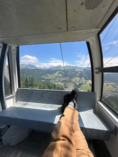 An interior view of a cable car cabin in Les Gets, France, depicts a person's lower body (legs and feet) resting on a light gray bench seat. The individual wears brown/tan pants, dark athletic shoes, and light blue-gray socks, with their legs crossed at the ankles. The cabin features light gray metal walls, a textured gray floor, and large panoramic windows offering a view of the mountainous landscape. The ceiling is composed of light gray riveted metal panels, with a circular speaker grill visible on the upper right. Outside, a vast expanse of green forested mountains and a village with numerous buildings are visible under a clear blue sky with scattered white clouds. Distant snow-capped peaks are present on the horizon. A dark cable, part of the lift system, runs vertically through the right side of the frame. The scene is illuminated by bright daylight, casting shadows on the bench.