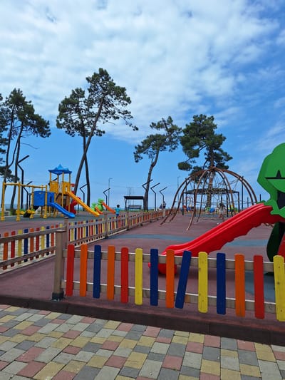 The image captures a vibrant children's playground situated in Kobuleti, Georgia. The scene is dominated by a colorful collection of playground equipment, including slides, climbing nets, and multi-colored fences. Tall, slender pine trees with dense green foliage frame the playground, their branches reaching towards a bright blue sky scattered with wispy white clouds. The time of day appears to be daytime, given the clear sky and the lighting. The weather seems pleasant, suggesting a good day for outdoor activities.

The playground features a large, geodesic dome-like climbing structure with netting, a bright red slide, and a smaller yellow slide. Various sections of the playground are delineated by fences made of brightly colored wooden slats in red, orange, yellow, and blue. The ground surface is a mix of reddish-brown rubberized material and patterned paving stones in shades of yellow, gray, and pink.

While the image is primarily focused on the playground, a few individuals are visible, suggesting the space is in use. A child can be seen in the distance interacting with the climbing structure, and another figure is standing near a swing set further back. These human elements add a sense of life and activity to the otherwise static playground structures. The overall impression is one of a cheerful and engaging recreational space set against a natural backdrop. There is no visible text in the image.