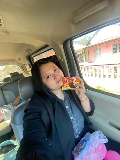 A woman wearing a black hijab and black jacket is sitting in the passenger seat of a car. She is holding a small package of "Better Caramel" cookies in her right hand. The woman is smiling and looking at the camera. Outside the car window, there are houses with red walls and tiled roofs, characteristic of Indonesian architecture. The interior of the car is visible, with grey seats and a light-colored ceiling. In the background, another person is partially visible in the backseat. The scene appears to be in the city of Jotangan, Indonesia, as indicated by the architecture and the presence of a motorcycle in the distance.
