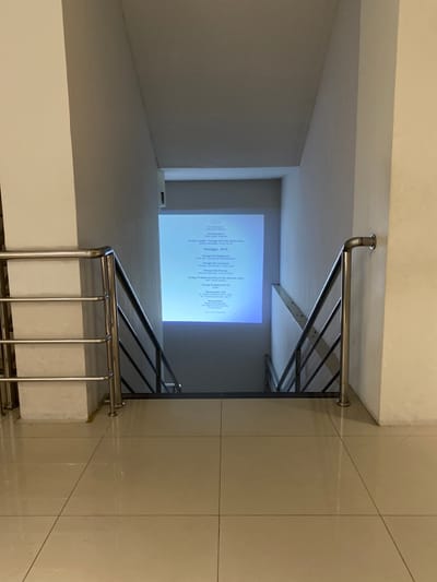 The image shows a view down a staircase in Banguntapan, Indonesia, located at coordinates -7.8 latitude and 110.407 longitude. There are stainless steel railings on both sides of the staircase, with a projected image of a document on a white wall at the end of the staircase. The document is titled "Tentagi Aji" and appears to be a list of items. The floor is tiled and there is a white wall to the right side of the frame. The stairs are made of concrete. The lighting is dim, casting a shadow across the bottom of the staircase. There are no people in the image.