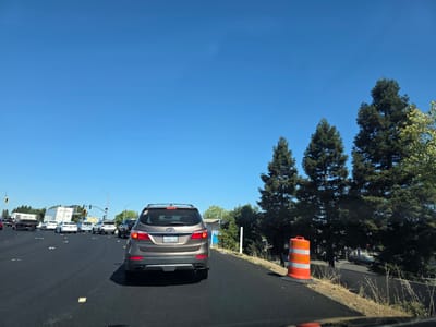 The image captures a view from the driver's seat of a car on a sunny day in West Sacramento, USA. The scene is a multi-lane road with moderate traffic. In the foreground, the rear of a brown SUV is visible, its license plate reading "8XNR210". The road surface is dark asphalt, with yellow rectangular markers.
Several other vehicles are on the road, including white cars, a white truck with a trailer, and a black SUV. In the distance, traffic lights and signs are visible. To the right of the road, there is a construction zone with an orange and white traffic barrel. Beyond that, a line of tall evergreen trees stands against a clear blue sky. The sun is bright, indicating it is likely daytime, and the clear sky suggests fair weather. The overall impression is one of a typical commute or travel on a highway. There is no discernible emotion conveyed by the image.
Visible text includes the license plate "8XNR210" on the SUV, and text on signs in the distance, though the text on the signs is illegible. A white trailer in the distance has text on its side, and a yellow sign also has text that is too blurry to read.
