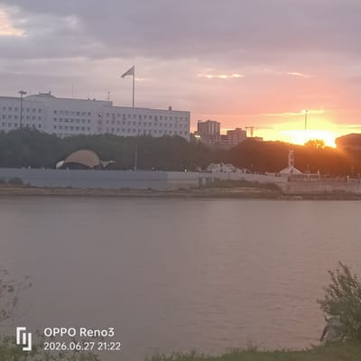 The image captures a sunset over the Ural River in Atyrau, Kazakhstan. In the foreground, the placid river occupies the lower half of the frame, reflecting the muted colors of the sky. The riverbank is visible on the right, with grass and a bush. A person is partially visible in the lower right corner, seemingly engaged in an activity near the water's edge.
Across the river, the Atyrau city skyline is depicted. A large, white, multi-story building with numerous windows dominates the left side of the cityscape. A flag pole with a white flag is prominent in front of this building. To the right of the building, a cluster of trees forms a green band. Further to the right, several other buildings of varying heights are visible, including some taller structures. A distinctive architectural feature, possibly a monument or sculpture with an arched top, stands on the riverfront promenade. In the distance, the setting sun casts a warm, golden glow across the sky, with clouds tinged with orange and pink. The overall atmosphere is serene, with the day's end marked by the soft light and tranquil water. A watermark at the bottom indicates the device used ("OPPO Reno3") and the date and time ("2026.06.27 21:22").