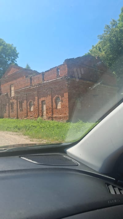 The image captures a dilapidated, red brick building with a partially collapsed roof, set against a clear blue sky and surrounded by green foliage. The building features arched window openings, some of which appear to be boarded up. The foreground is dominated by the interior of a vehicle, specifically the dashboard with a visible speaker grille and an "AIRBAG" indicator. The scene suggests an outdoor view from inside a car, possibly on a sunny day. The location is identified as Дзяржынскі сельскі Савет, Belarus.