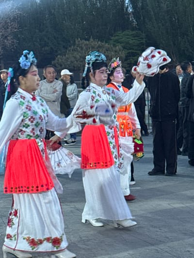Two pairs of elaborately dressed individuals in traditional Chinese attire, including white gowns with floral patterns and red accents, are posing outdoors. The performers have ornate headwear and makeup, typical of Chinese opera or folk dance. In the background, blurred figures of onlookers are visible on what appears to be a stone or concrete pavement. The setting suggests an urban environment, possibly a park or public space. The picture indicates that it's a cultural performance or street festival in Jinzhou, China.