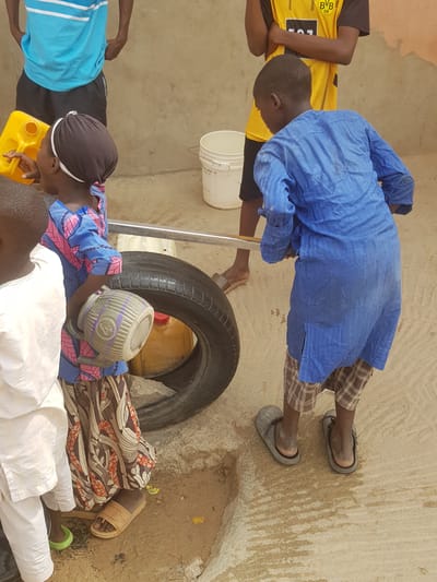 The image depicts a group of people outdoors in what appears to be a semi-urban area, potentially in Dutsinma B, Nigeria. Several individuals are gathered around a tire and various containers, possibly involved in a water-related activity. One person, wearing a blue garment and plaid shorts, leans over a metal pole positioned near a white bucket. Another person, wearing a blue and pink patterned shirt, rests a plastic pitcher on a white garment. Other people are visible wearing different colored shirts. The environment consists of a concrete surface and what looks like a wall in the background.