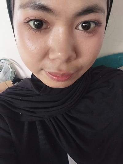 The image is a close-up selfie of a young woman wearing a black hijab and a black top. She is indoors, in what appears to be a room with a white wall and a teal-colored object in the background. The woman has dark eyes, dark eyebrows, and light-colored skin. Her lips are painted with a glossy, pinkish-red lipstick. There are some items visible in the background to the left, including what appears to be a plastic bag with white and green material inside, and a cardboard object. The lighting suggests an indoor setting, possibly natural light coming from the side, as evidenced by the highlight on her cheek. The specific location in the city of Jeppee, Indonesia, cannot be determined from the image alone.