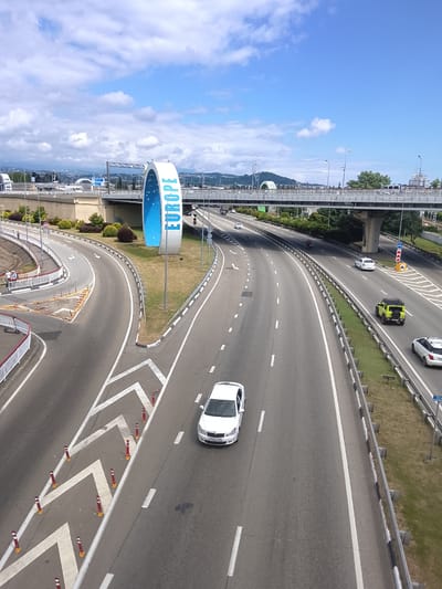 This media file depicts a view of a multi-lane highway under a bright, partly cloudy sky, suggesting daytime. The scene is set on a wide, paved roadway with clear lane markings. A prominent architectural element, a large, blue, arch-like structure with the word "EUROPE" written vertically in white letters, stands to the left of the highway.

Several cars are visible on the highway, including a white sedan in the foreground, a yellow SUV, and other vehicles further down the road. The highway is bordered by guardrails and grassy verges. In the background, an elevated pedestrian bridge crosses the highway, and beyond that, hills and buildings are visible. The overall impression is one of an open, modern transportation hub, possibly a border crossing or a significant junction. The weather appears pleasant, with the sun shining through scattered clouds. There are no discernible people actively engaged in an event, though a few individuals are seen near the "EUROPE" structure and on the side of the road. The location cues, such as the "EUROPE" sign, suggest a place of geographical significance.