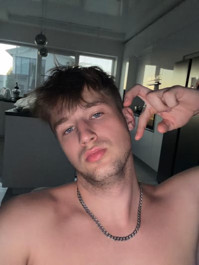 The image is a selfie taken by a shirtless young man with light brown, messy hair and blue eyes. He is wearing a silver chain necklace. His right hand is raised, with his index and middle fingers bent, positioned near his ear. He has a light beard and his lips are slightly parted. The background appears to be an indoor setting, possibly a kitchen or living area, with large windows showing an exterior view of a building and trees. The lighting is dim, suggesting it might be evening or early morning. There are no discernible details that would place the location in Vanino, Russia.