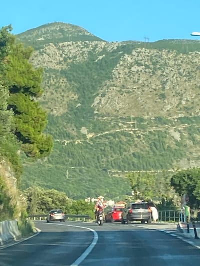 In Soline, Croatia, a road curves through a mountainous, vegetated landscape under a clear blue sky. The foreground shows a paved road with white lane markings and concrete barriers on the left. Several vehicles are present on the road, including a dark-colored SUV, a red car, and a motorcycle with a rider wearing a helmet and orange clothing. The motorcycle is positioned behind the red car, and the SUV is further ahead. A triangular caution sign with an exclamation mark is visible to the right of the motorcycle. In the background, a steep mountain slope is covered with dense green trees and rocky outcrops. Power lines and structures are visible on the mountain's upper reaches. Lush green trees, including a large pine tree, line the left side of the road in the mid-ground.
