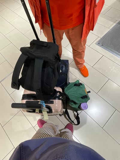 This image presents a top-down view of two individuals and their accumulated luggage on a tiled floor in Crawley, United Kingdom.
One individual, positioned further away, is observed from the waist down, wearing an orange long-sleeved shirt, an unbuttoned orange jacket, and peach-colored trousers. One foot is visible, encased in a bright orange loafer with a gold-colored chain across the vamp.
The second individual, closer to the camera, is also viewed from the waist down, wearing striped trousers (brown and white) and a dark blue upper garment. One foot is visible, clad in a dark pink loafer.
The luggage collection consists of:
1. A black rolling suitcase with its two-part extendable handle visible, extending upwards between the two individuals.
2. A black backpack placed directly on top of the black rolling suitcase.
3. A light pink rigid-shell carry-on suitcase, featuring a black telescoping handle and an attached blue identification tag, positioned next to the black luggage.
4. A navy blue bag with a textured surface, located between the black backpack and the pink suitcase.
5. A green soft-sided bag, possibly a small backpack or pouch, resting on the floor beside the pink suitcase.
6. A purple container, consistent with a bottle or thermos, situated next to the green bag.
The floor is composed of light-toned square tiles separated by dark grout lines. A rectangular floor mat with a metallic frame is visible on the right side of the image. The environment suggests an indoor public space.