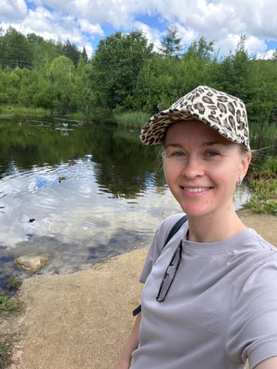 A woman is taking a selfie in front of a lake on a cloudy day. She is wearing a grey t-shirt and a leopard-print baseball cap. She is smiling at the camera. There are trees and foliage surrounding the lake, with some ducks swimming in the water. The woman is standing on a sandy shore.