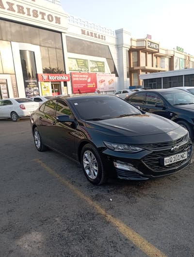 A black Chevrolet Malibu is parked in a lot outside a shopping complex in Termez, Uzbekistan. The building features large signage with "AXRISTON" and "MARKAZI" prominently displayed. Several other vehicles, including a white sedan, are also parked in the lot. The ground is asphalt with yellow parking lines. The sky is clear and appears to be late afternoon or early evening.