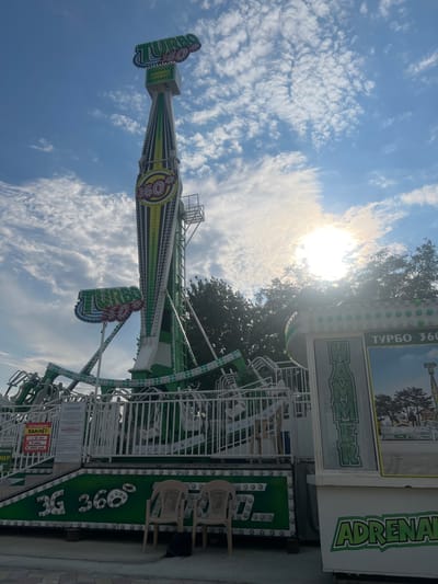 An amusement park ride, identified as "Turbo 360 Hammer," is the central subject of this outdoor photograph, taken during daylight hours in Golubitskaya, Russia. The ride is a towering, multi-colored structure with green, white, and yellow accents, featuring a large vertical arm that swings riders. The sky above is blue with scattered white clouds, and the sun is partially obscured by clouds, casting a bright glare. The base of the ride is a platform with white railings and signage. One sign displays "ATTRAKTION HAMMER" in Russian, along with age and price restrictions. Below the ride's structure, the words "3G 360°" are emblazoned in large green letters. Two empty beige plastic chairs and a dark backpack are placed on the ground in front of the ride. The scene appears to be set within an amusement park or fairground, with trees visible in the background.