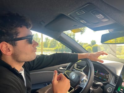 The image displays a male individual seated in the driver's position of a modern Renault vehicle, located in Orléans, France. The male, observed from the chest up, has dark, curly hair and wears dark Ray-Ban sunglasses, a dark zip-up jacket over a white t-shirt, and a silver bracelet on his left wrist. He is oriented towards the right, looking forward and slightly outwards. His right hand rests on the lower spoke of the steering wheel with the thumb raised, while his left hand is lightly placed on the dashboard directly above the digital instrument cluster. The instrument cluster indicates "0 km/h" and displays a green battery charge icon. An SOS button is visible on the overhead console. The scene outside the vehicle is brightly lit by sunlight, revealing a body of water lined with green trees and foliage in the mid-ground, reflecting sunlight. The car appears to be stationary or moving very slowly.