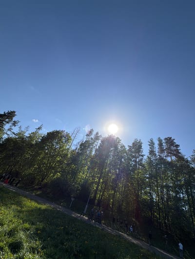 The image shows a sunny day in a park near Oreshniki, Russia. The bright sun is high in the sky, partially obscured by trees on the right. The trees are tall and green, lining a path visible on the right side. People are present on and beside the path, suggesting a gathering or event. The ground slopes slightly downward from right to left. The sky is a clear blue.