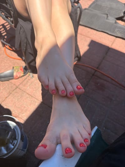 The image shows a close-up view of a person's feet, which have been painted with bright red nail polish. The feet are resting on a tiled outdoor surface, likely a patio or walkway, made of reddish-brown tiles. Sunlight casts distinct shadows across the scene, suggesting it is daytime.

One foot is positioned in the foreground, with its toes clearly visible and the red polish shining. The other foot is slightly behind and to the left, with its leg visible extending upwards. The person appears to be outdoors, possibly relaxing. There are some objects in the background, including a black mat with a textured surface, a thin orange cord, and a dark green object that might be part of a piece of furniture. A metallic container, possibly a pot or bucket, is visible in the lower right corner of the image.

The overall impression is one of casual relaxation in a sunny outdoor environment. The bright nail polish adds a pop of color to the scene. No text is visible in the image.