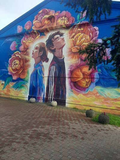 This is a medium shot of a mural on the side of a building in Shelekhov, Russia. The mural depicts a young man and woman with their eyes closed, looking upwards, surrounded by large, colorful flowers. The man is wearing a dark jacket with white stripes, and the woman is wearing a blue shirt. The background is a vibrant blue sky, transitioning to an orange and yellow sky at the bottom. In the foreground, there is a red brick pavement with some wet patches, and two large, round planters with small red flowers. A dark green tree branch partially obscures the right side of the mural. The overall scene suggests an artistic and somewhat serene outdoor environment.